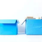 Siisti sininen muovilaatikko ja rypistynyt pahvilaatikko vierekkäin vertailussa. Muovilaatikossa tavarat järjestyksessä, pahvilaatikossa sekaisin. Valkoinen tausta, sinitehosteet.