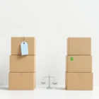 Muuttolaatikot vertailussa: vasemmalla sininen vuokraushintamerkki, oikealla vihreä omistussymboli, välissä kallistuva vaaka. Minimalistinen sommitelma valkoisella taustalla.